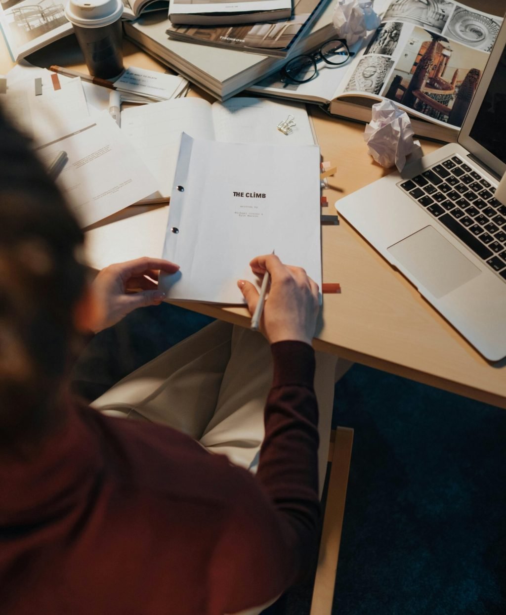 A person writing notes on a screenplay surrounded by books and a laptop on a work desk.