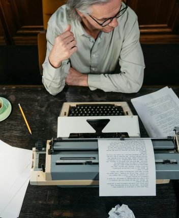 An overhead shot of an adult man writing with a typewriter in a vintage-themed study.
